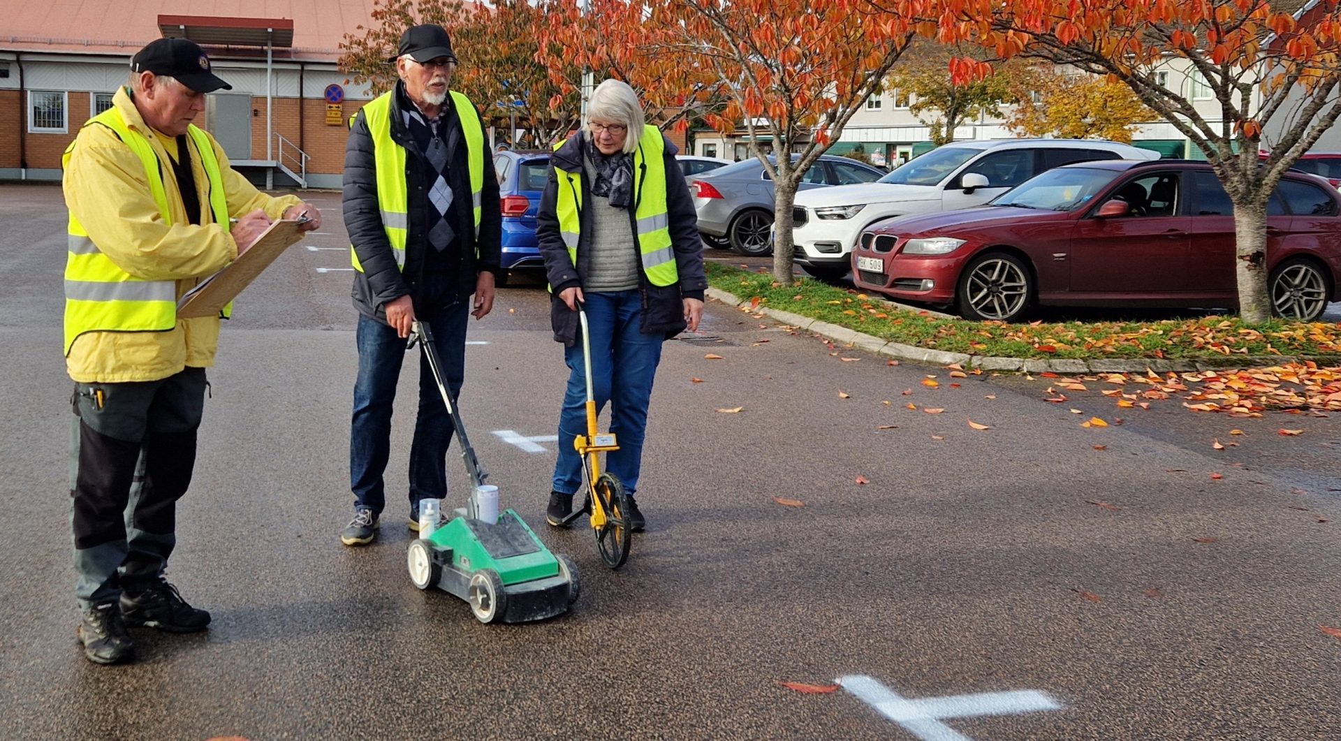 Tre personer står på en parkeringsplats och arbetar. De bär gula reflexvästar och markerar rutor på marken. En person skriver på ett klippbord, medan de andra två använder mät- och markeringsverktyg på marken. I bakgrunden syns parkerade bilar och träd med höstfärgade löv.