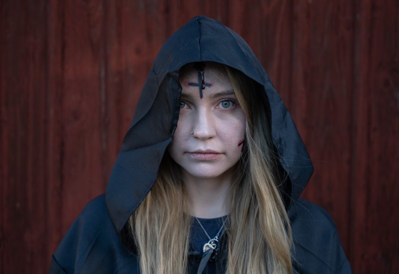 Lo Elmqvist står framför ett rött hus. Hon bär en svar mantel med huva. Lo är vitsminkad i ansitet och har ett upp-och nedvänt blodigt kors i pannan