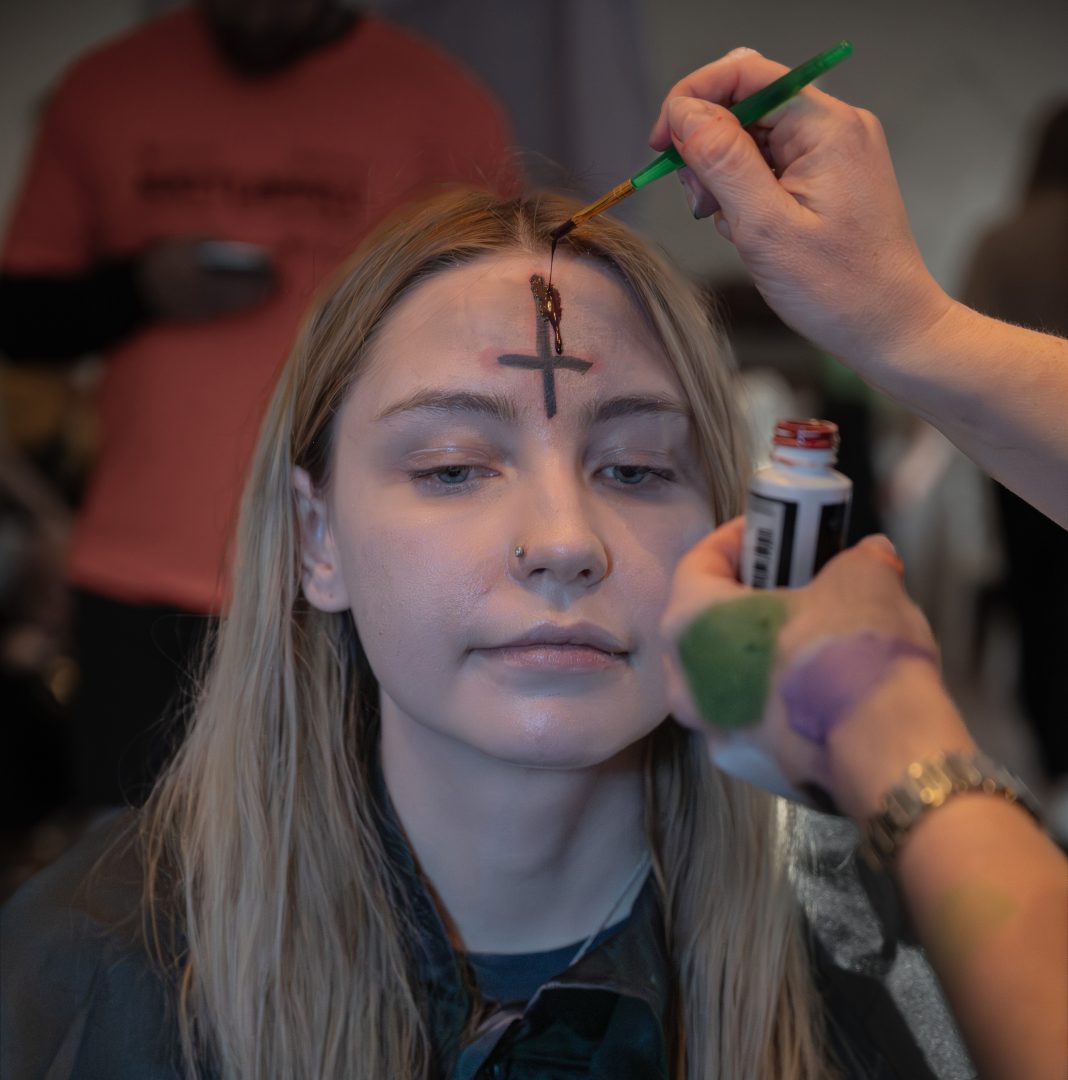 Lo Elmqvist är centrerad i bilden, hon är vitmålad i ansiktet och har ett svart, upp- och nedvänt kors i pannan. Från högra hörnet sticker Martina Danielssons armar fram. I ena handen håller hon en flaska med låsasblod, i den andra håller hon en pensel. Penseln har hon doppat i flaskan. Med penseln målar hon låsasblod på korset i Los ansikte.