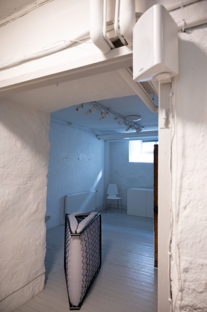 På bilden syns en dörröppning som leder till ett släckt rum. Rummet är vitt och har stenväggar. I taket sitter det spotlights. På golvet står en ihopvikt tältsäng. Längst in står en vit stol. På väggen längst bak sitter ett fönster i höjd med taket.