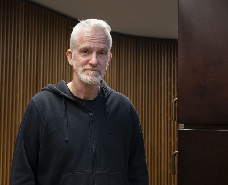 En äldre man med kort vitt hår och skägg står inomhus framför en vägg med vertikala trälister. Han bär en svart huvtröja och tittar mot kameran med ett neutralt uttryck.