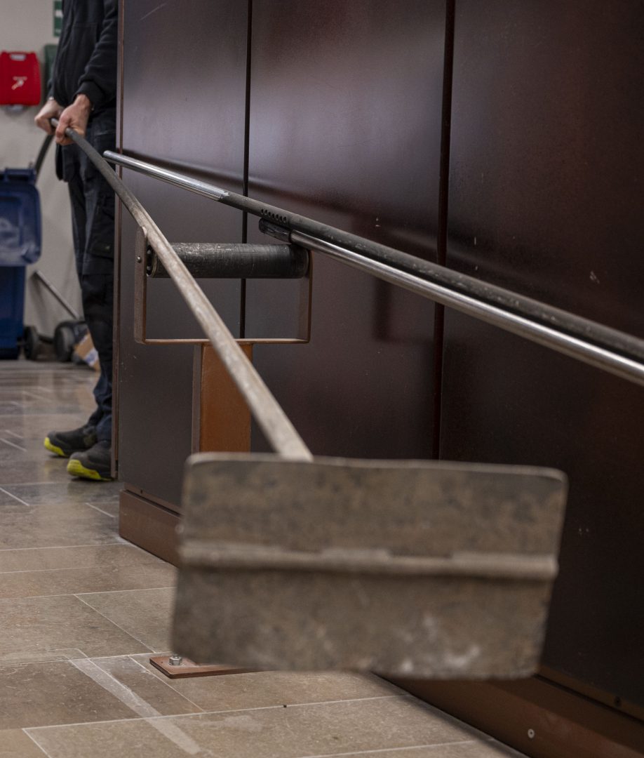 En person håller i ett långt metallverktyg med en bred, platt ände som vilar mot ett stativ. Verktyget används inomhus i en lokal med stengolv och mörka paneler.