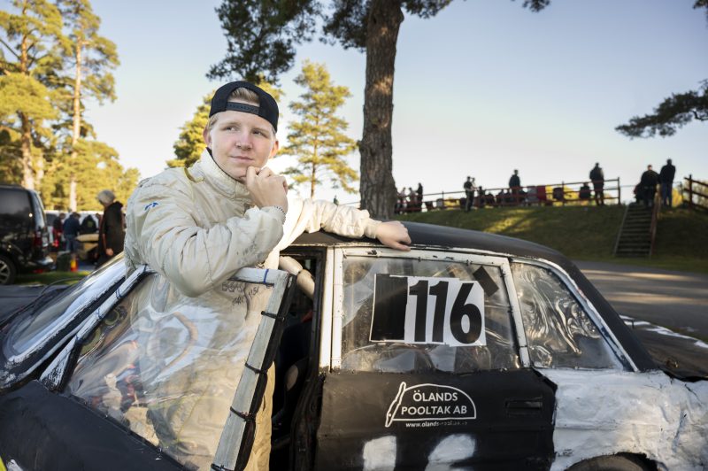 En kille i ljus föraroverall och bakåtvänd keps står framför sin svartvita folkracebil med startnummer 116 och ser fundersam ut.