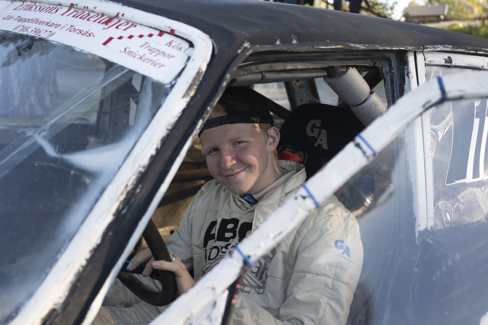En ung man i vit racingoverall och bakåtvänd keps sitter i en tävlingsbil och håller i ratten. Han tittar mot kameran och ler. Bilen har ett enkelt, hemmabyggt utseende med skyddsbåge och plastfönster. Startnummer 116 syns delvis på rutan.