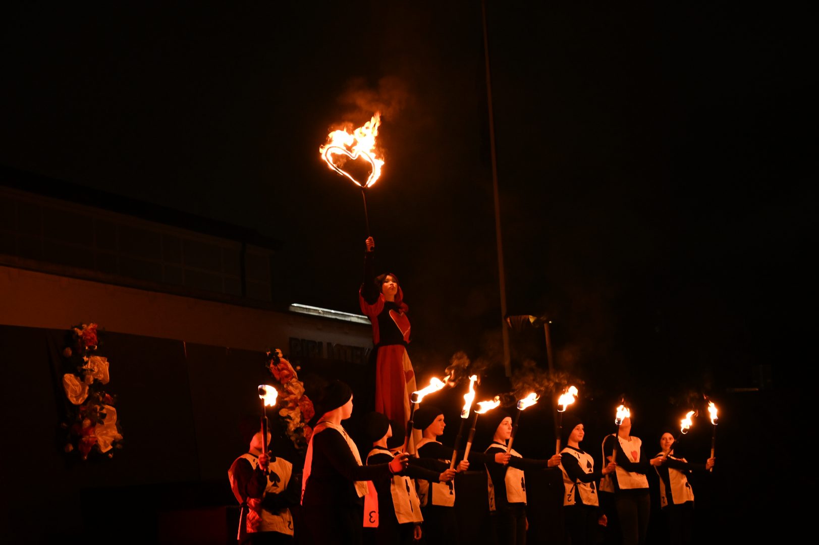 En person klädd som den röda drottningen ur Alice i Underlandet står upphöjd och håller ett brinnande hjärta, omgiven av figurer med facklor
