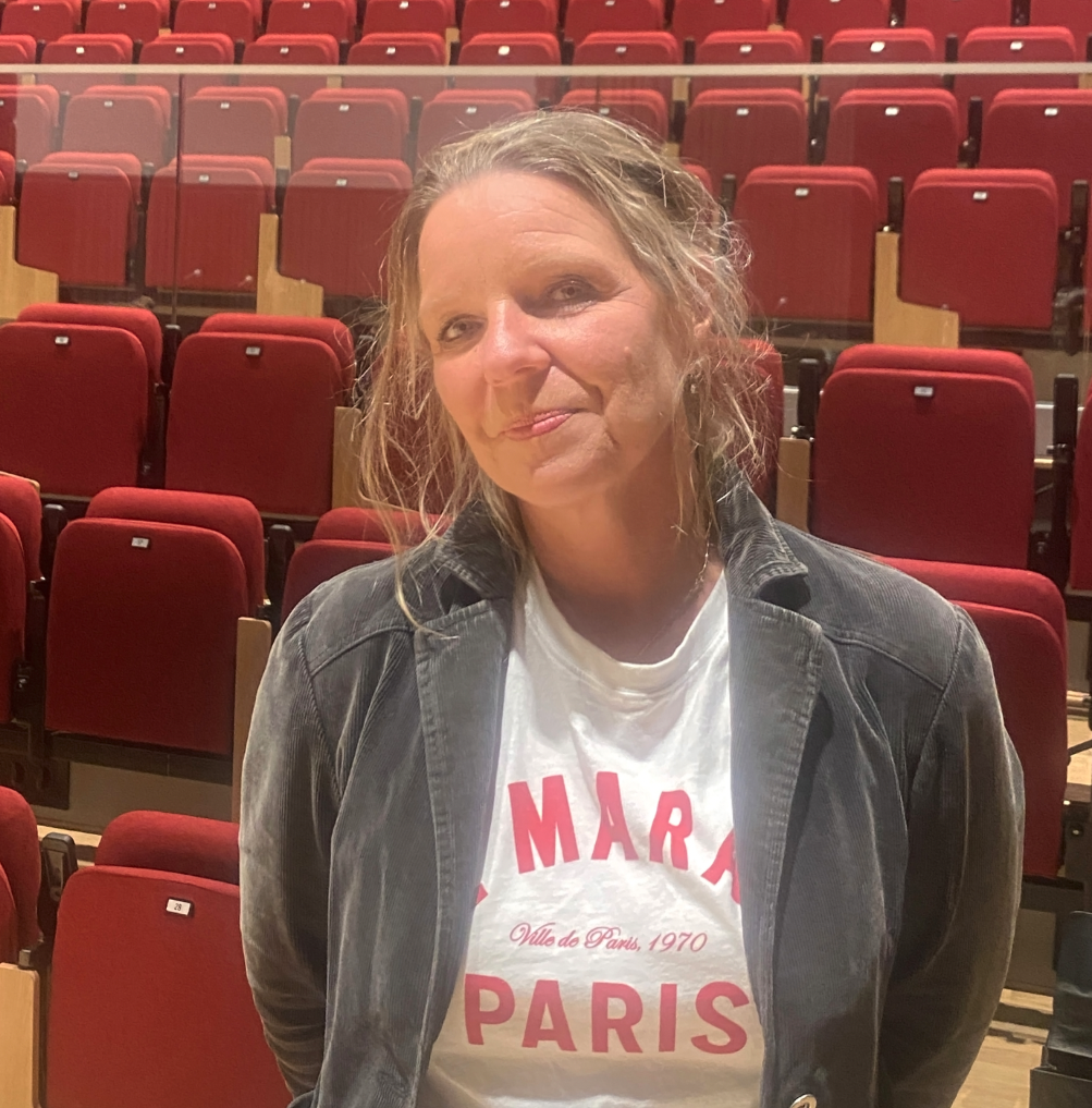 En kvinna står inomhus framför en rad röda teaterstolar. Hon har blont hår som är uppsatt lite slarvigt, med några slingor som hänger löst kring ansiktet. Hon ler svagt mot kameran. Hon bär en vit T-shirt med röd text, samt en mörkgrå jacka ovanpå.