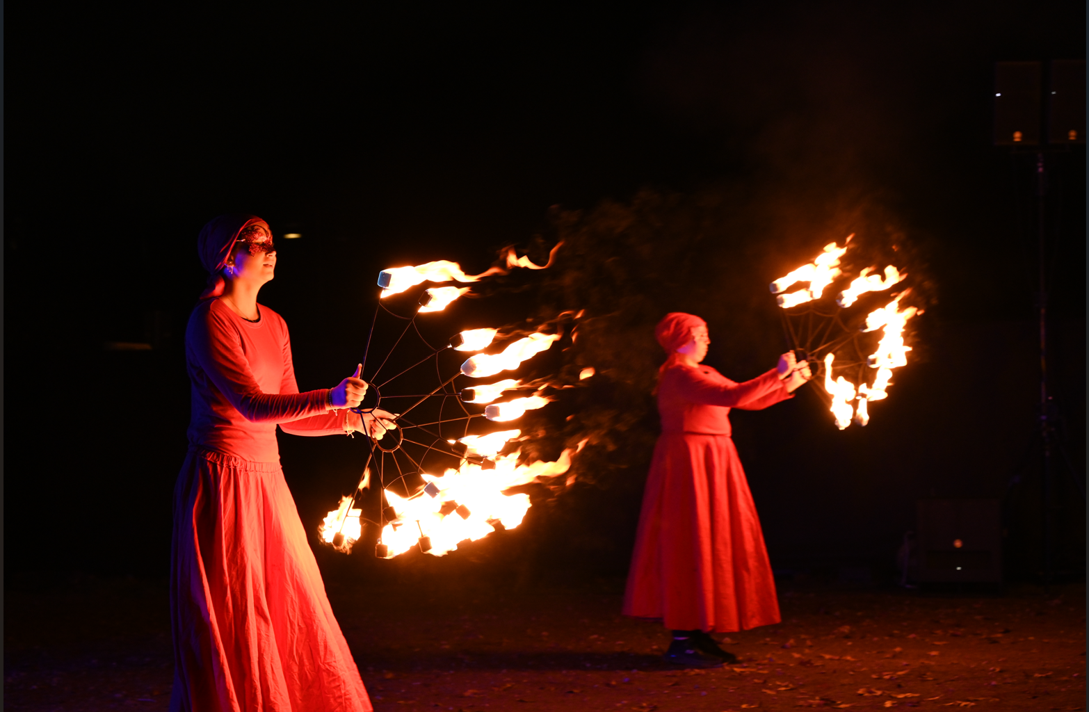 Två personer uppträder i mörkret med eldrekvisita. De bär långa, röda kläder och huvuddukar. Var och en håller i ett redskap med flera metallpinnar som har brinnande lågor i ändarna. Elden lyser starkt mot den mörka bakgrunden och speglar sig i deras kläder. Marken är täckt av löv, vilket antyder att det är utomhus under hösten.
