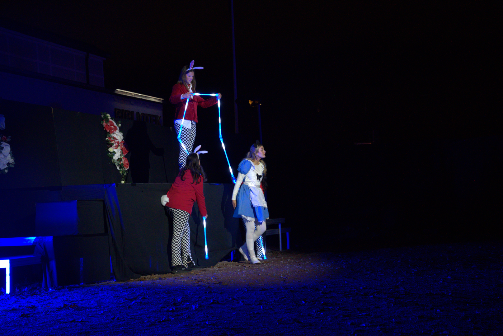 En utomhusscen i mörker, upplyst av blått ljus. Tre personer uppträder i kostymer. Två av dem är klädda som kaniner med röda jackor, rutiga byxor och vita svansar, varav en står på en upphöjd plattform och håller ett lysande blått rep. Den tredje personen, klädd i en blå klänning med vit förkläde och hårband utklädd till Alice, står nedanför och tittar upp mot dem. Scenen är dekorerad med konstgjorda blommor, och marken framför är täckt av löv.