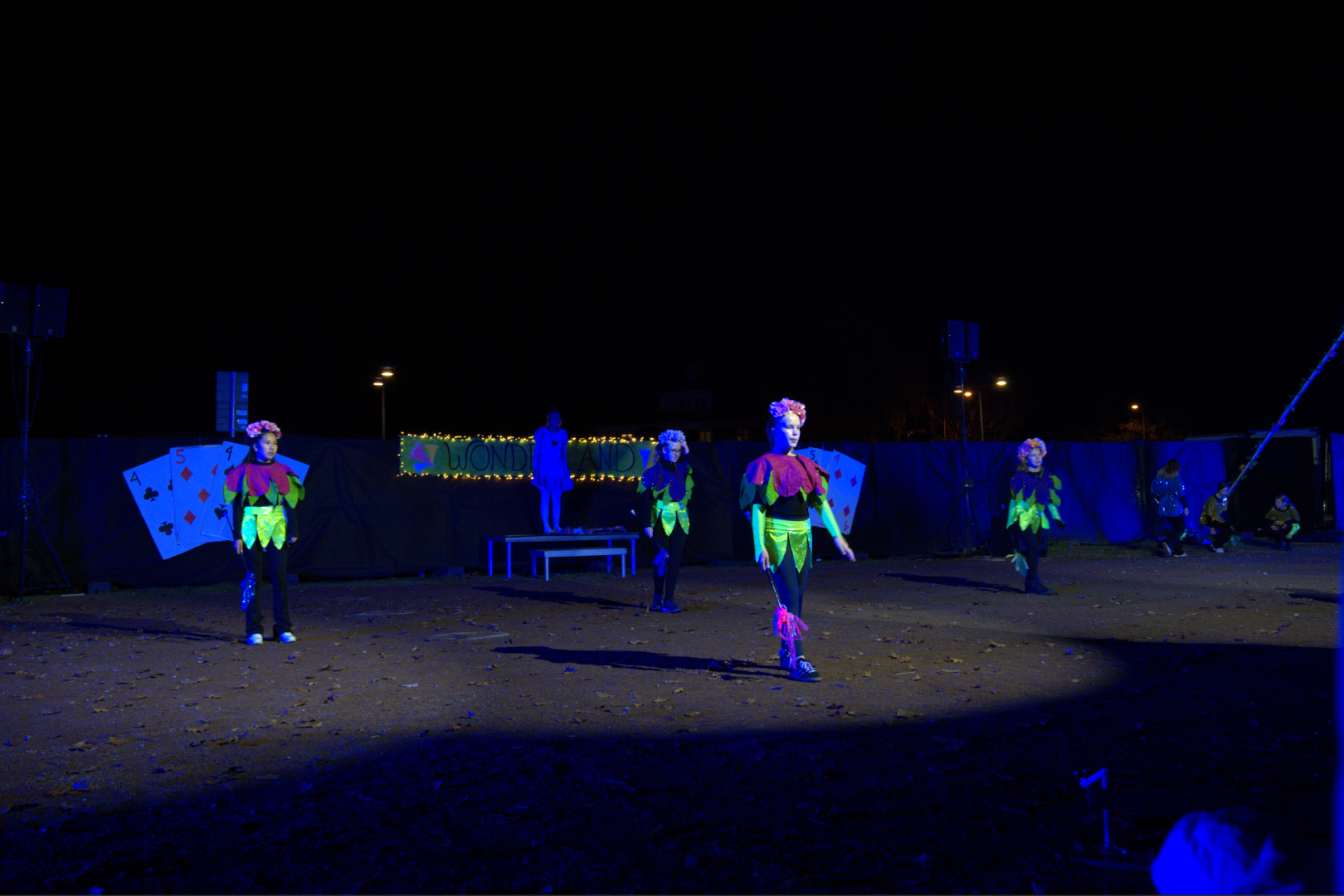 Fyra barn klädda som blommor uppträder i mörkret framför Alice i underlandet scenen med drill pinnar i handen av olika färger.