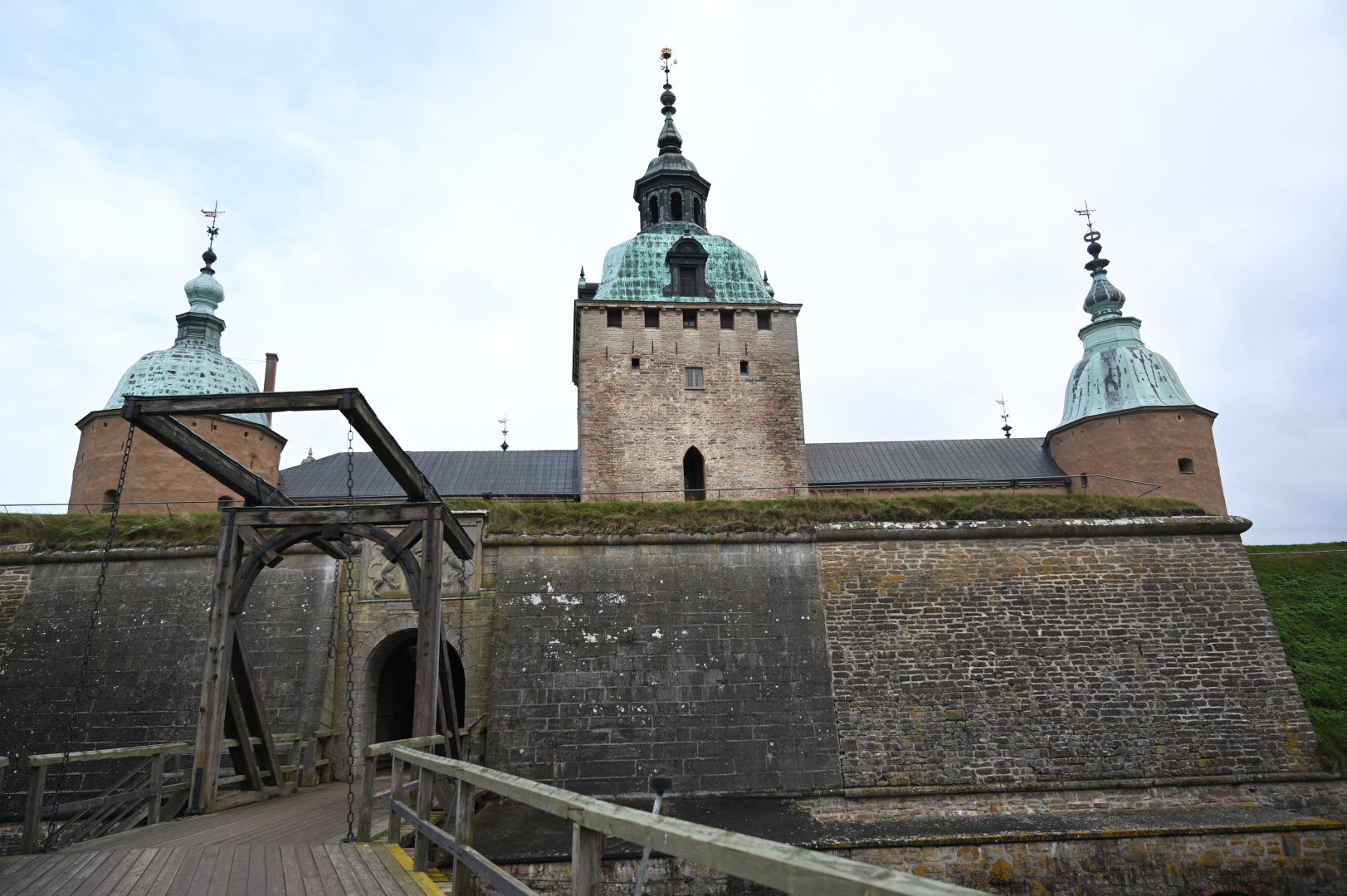 Bild på Kalmar slott sedd från vallgraven med den öppna träbron i förgrunden. Slottets robusta stenmur och tre torn med kopparklädda tak syns tydligt under en molnig himmel.