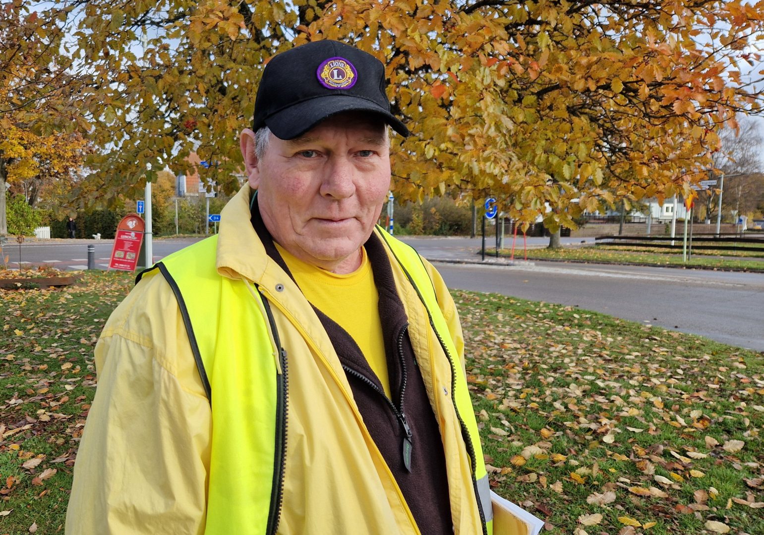 En äldre man står utomhus på en trottoar. Han bär en gul reflexväst ovanpå en gul tröja och en mörk jacka. På huvudet har han en svart keps med ett märke som visar Lions Clubs logotyp. Bakom honom syns träd med gula och orangea höstlöv, en väg och några trafikskyltar. Marken är täckt av fallna löv. 