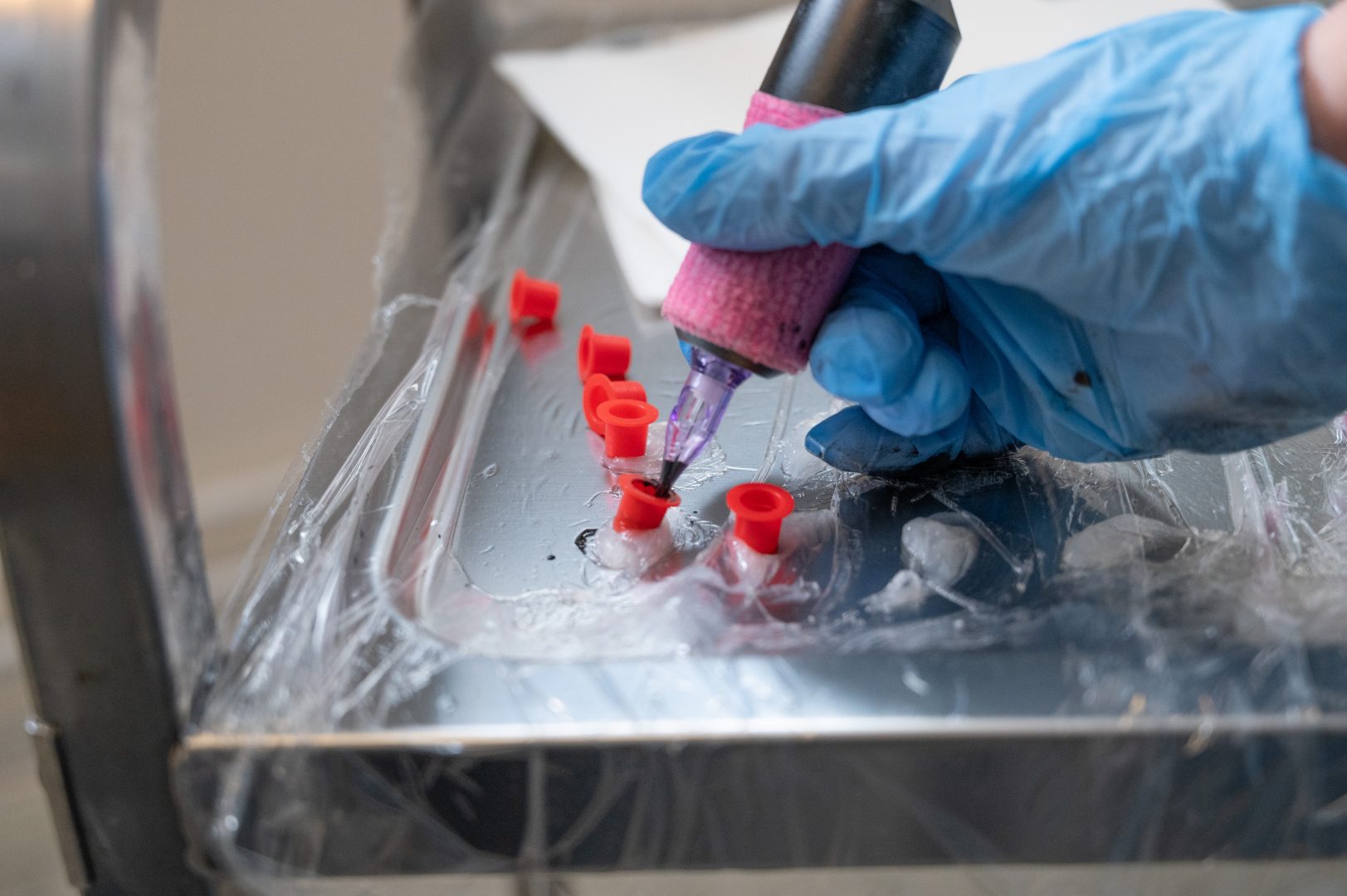 En närbild visar en hand med blå engångshandske som håller i ett tatueringsverktyg. Verktyget doppas i svart färg som finns i små röda färgkoppar placerade på en metallbricka täckt med plast. 
