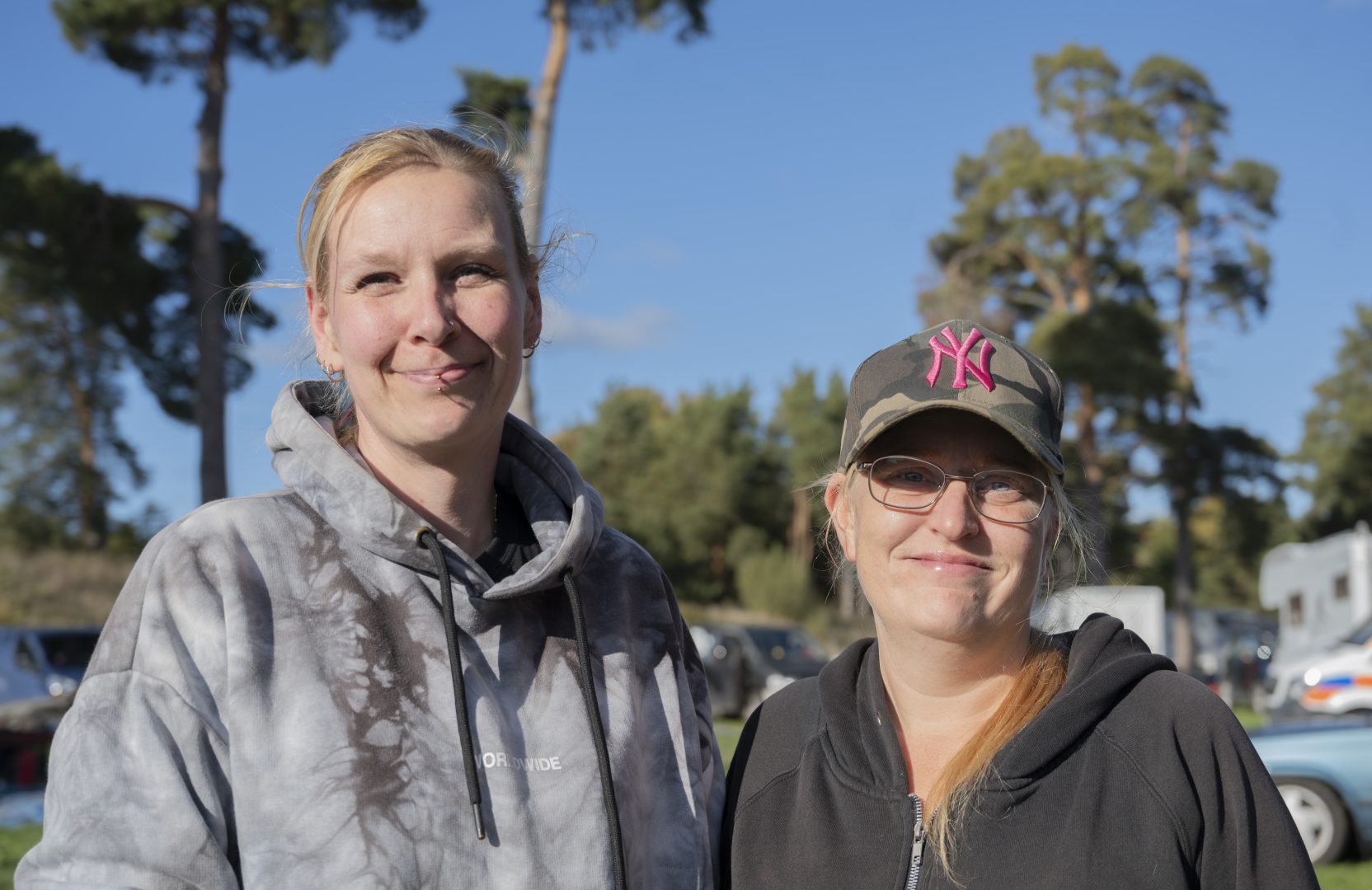 Två kvinnor står utomhus i soligt väder med träd och bilar i bakgrunden. Den ena bär en grå hoodie och har en piercing i läppen, den andra har glasögon, en mörk tröja och en kamouflagefärgad keps med rosa NY-logga. Båda ler mot kameran. 