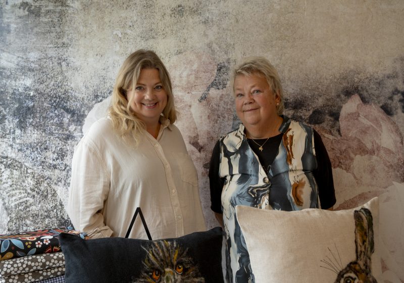 Två kvinnor står bredvid varandra framför en mönstrad vägg. Framför dem ligger textilier och kuddar med djurmotiv.