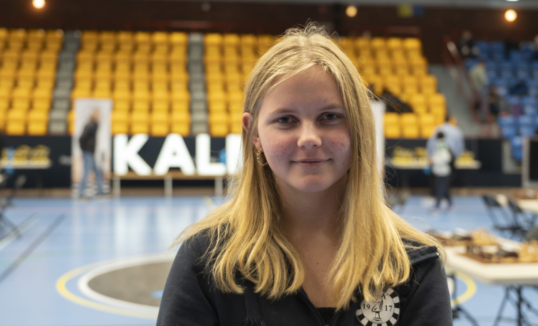 En ung blond flicka i 15 års åldern med som står i en idrottshall med läktare i bakgrunden