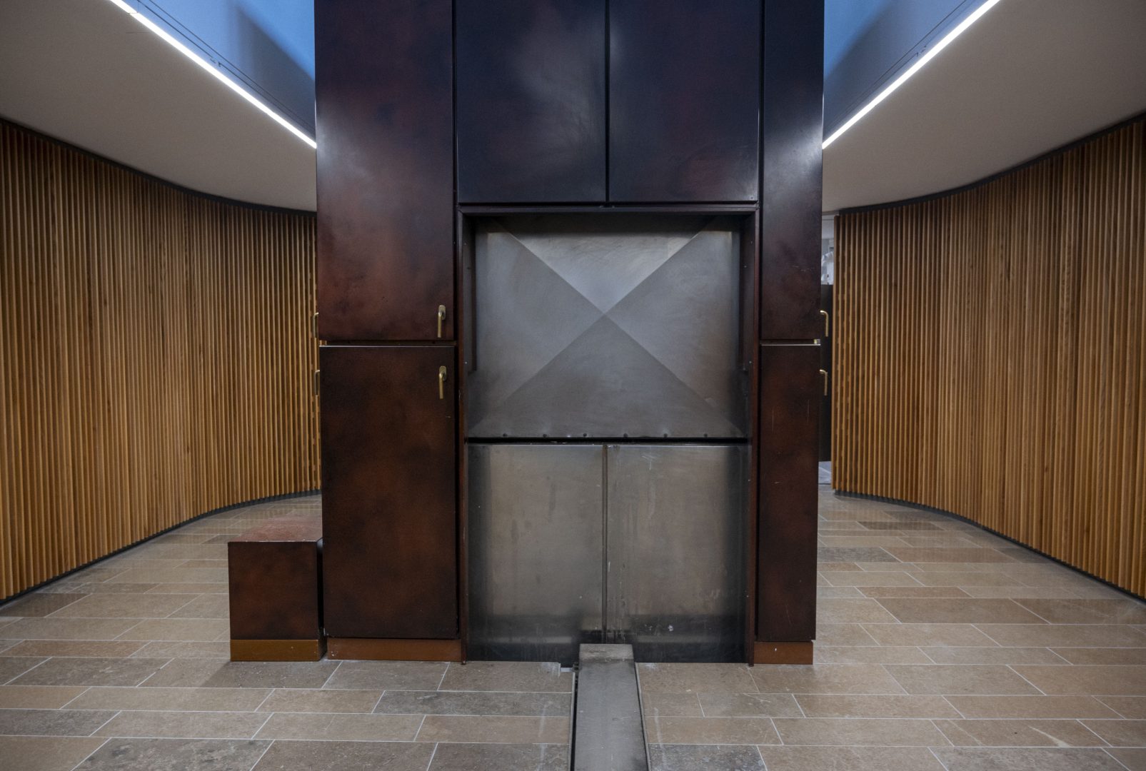 En stor metallugn med mörkbruna paneler och mässingshandtag står i ett rum med stengolv och väggar klädda i vertikala trälister. En metallplanka i golvet löper ut från ugnens nedre del. 