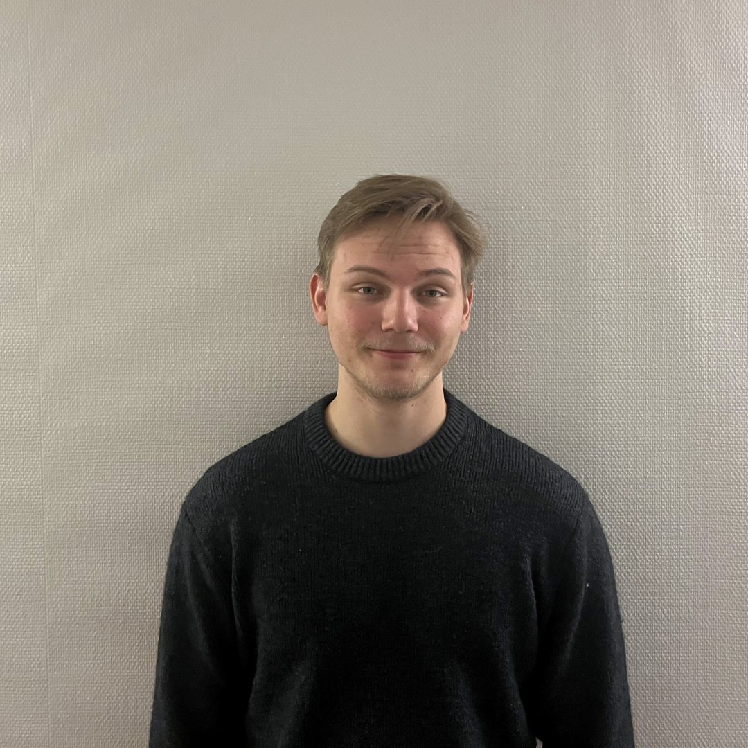 En ung man står framför en ljusgrå, lätt strukturerad vägg. Han har kort, ljusbrunt hår och bär en mörk stickad tröja. Han ser rakt mot kameran med ett svagt leende och har armarna avslappnat längs sidorna. Ljuset är mjukt och jämnt fördelat i rummet.