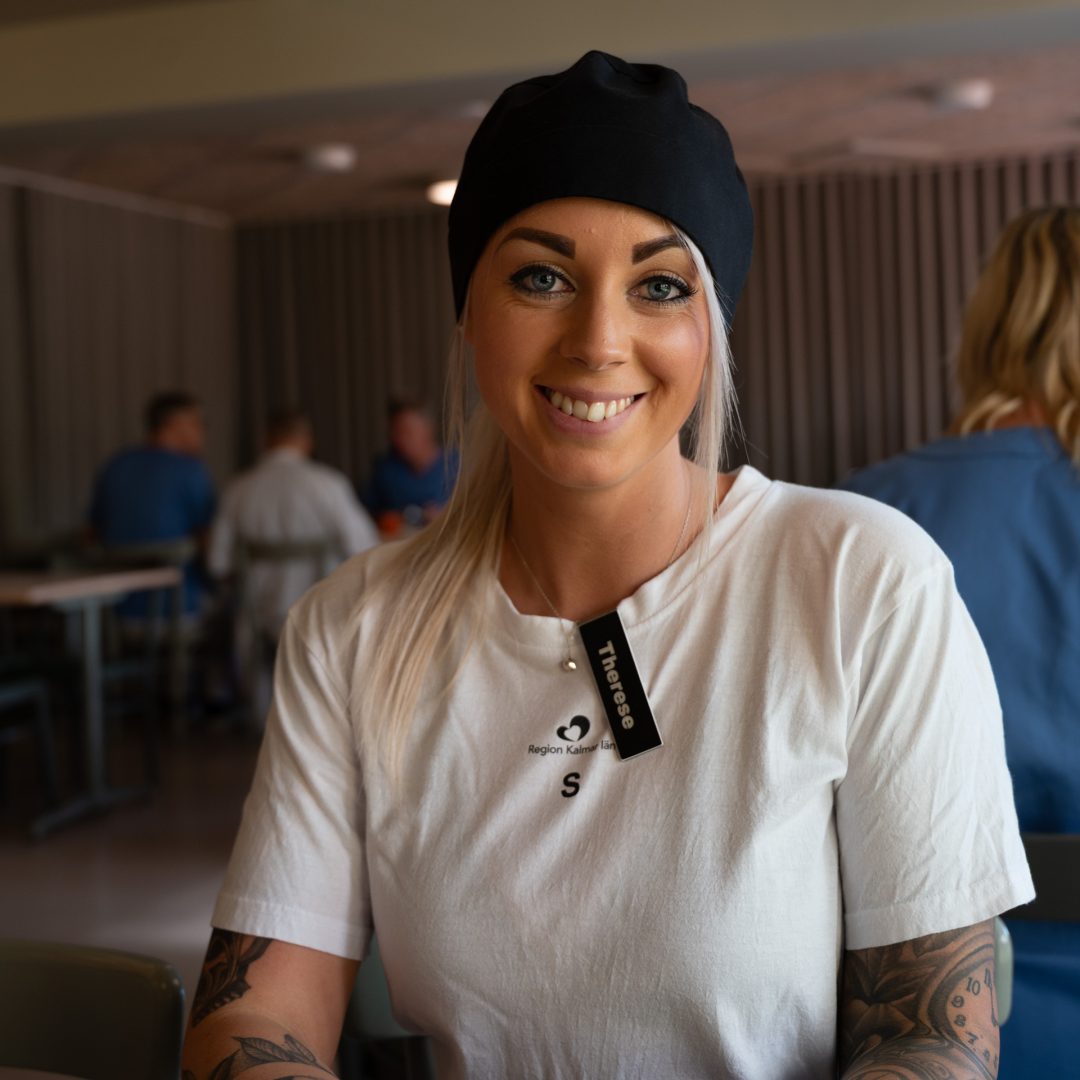 Therese, restaurangansvarig på Kalmar länssjukhus, sitter leende i matsalen iförd arbetskläder med Region Kalmar läns logotyp och namnbricka.