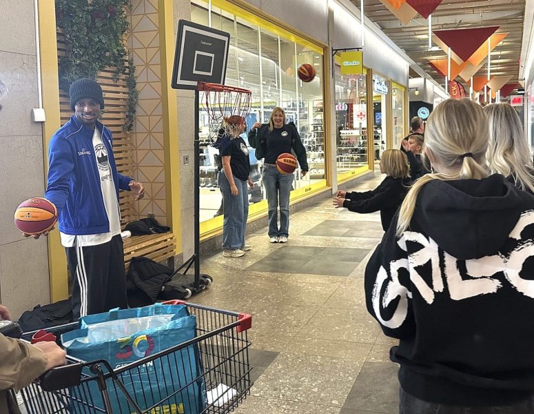 Folk står på kö. Det finns en basketkorg. En man håller i en basketboll. Två basketbollar flyger i luften. I vänstra hörnet kan man se en kundvagn.