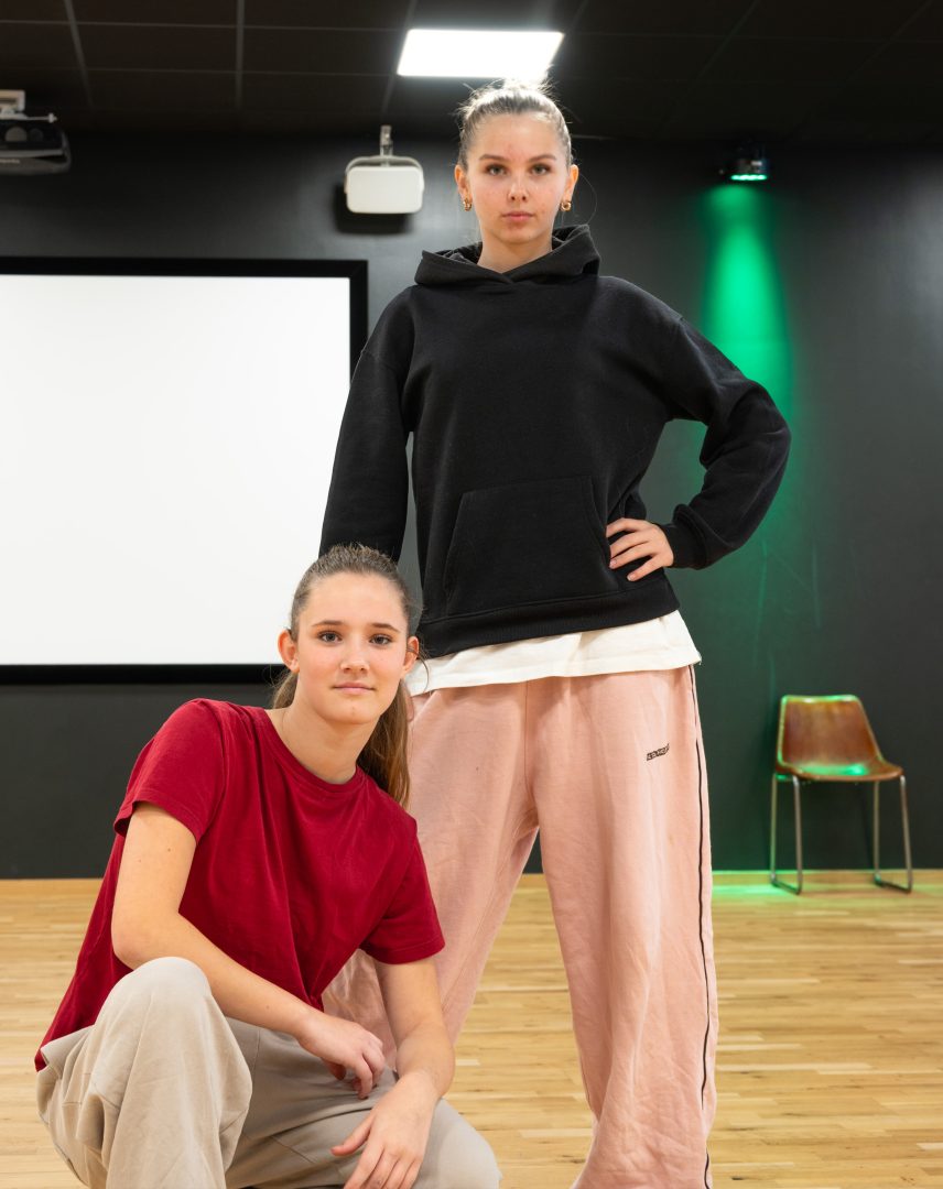 två tjejer som står mitten i danssalen en av tjejen till vänster hukar sig ner och poserar för kameran hon har röd tröja och grå byxor. en andra tjejen står upp till höger om den andra tjejen hon poserar framför kameran och har på sig en svart tröja och rosa byxor 