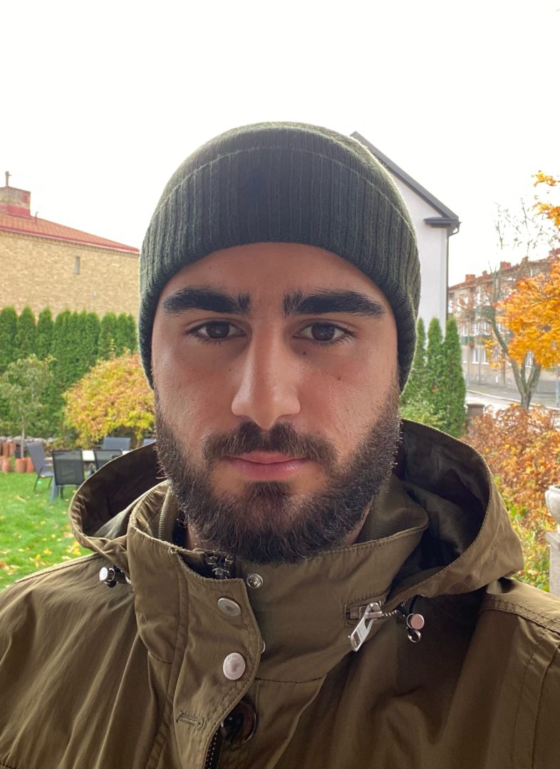 Bilden visar en Noel Bethgazo som står utomhus. Han bär en mörkgrön stickad mössa och en jacka i en liknande grön nyans med uppfälld krage. Personen har mörkt skägg och tittar rakt in i kameran med neutral min. I bakgrunden syns en trädgård med grönt gräs, stolar och bord, samt buskar och träd i höstfärger – gulorange och rött.