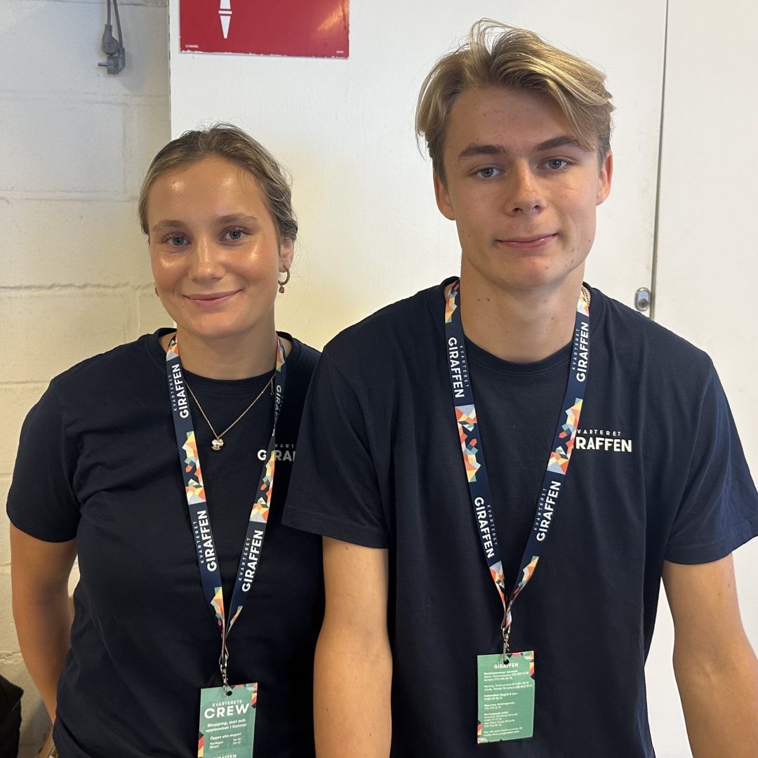 En tjej och en kille ler och tittar in i kameran. De bär likadana blå t-shirts och passerkort runt halsen . De har båda ljust hår.