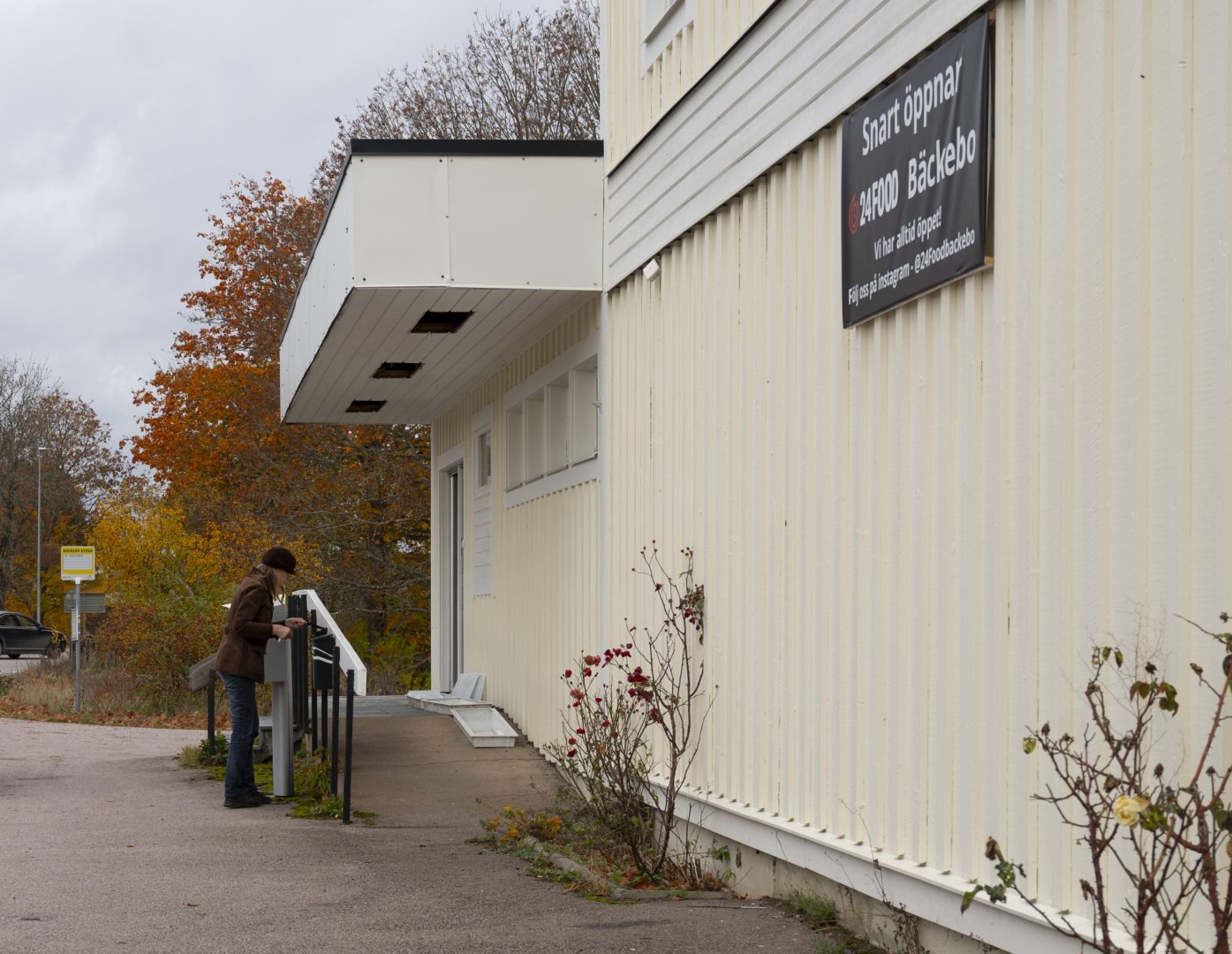 En kvinna lägger ner något i en brevlåda bredvid ett stort gult hus. På huset sitter en skylt om att det snart ska öppna en butik där. I bakgrunden syns träd som har orangea och bruna löv.