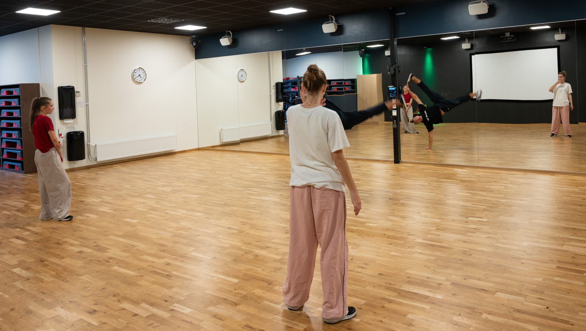 En grupp personer tränar dans i en spegelsal med trägolv. en person utför en akrobatiskt danssteg på en hand medan den andra tittar på 