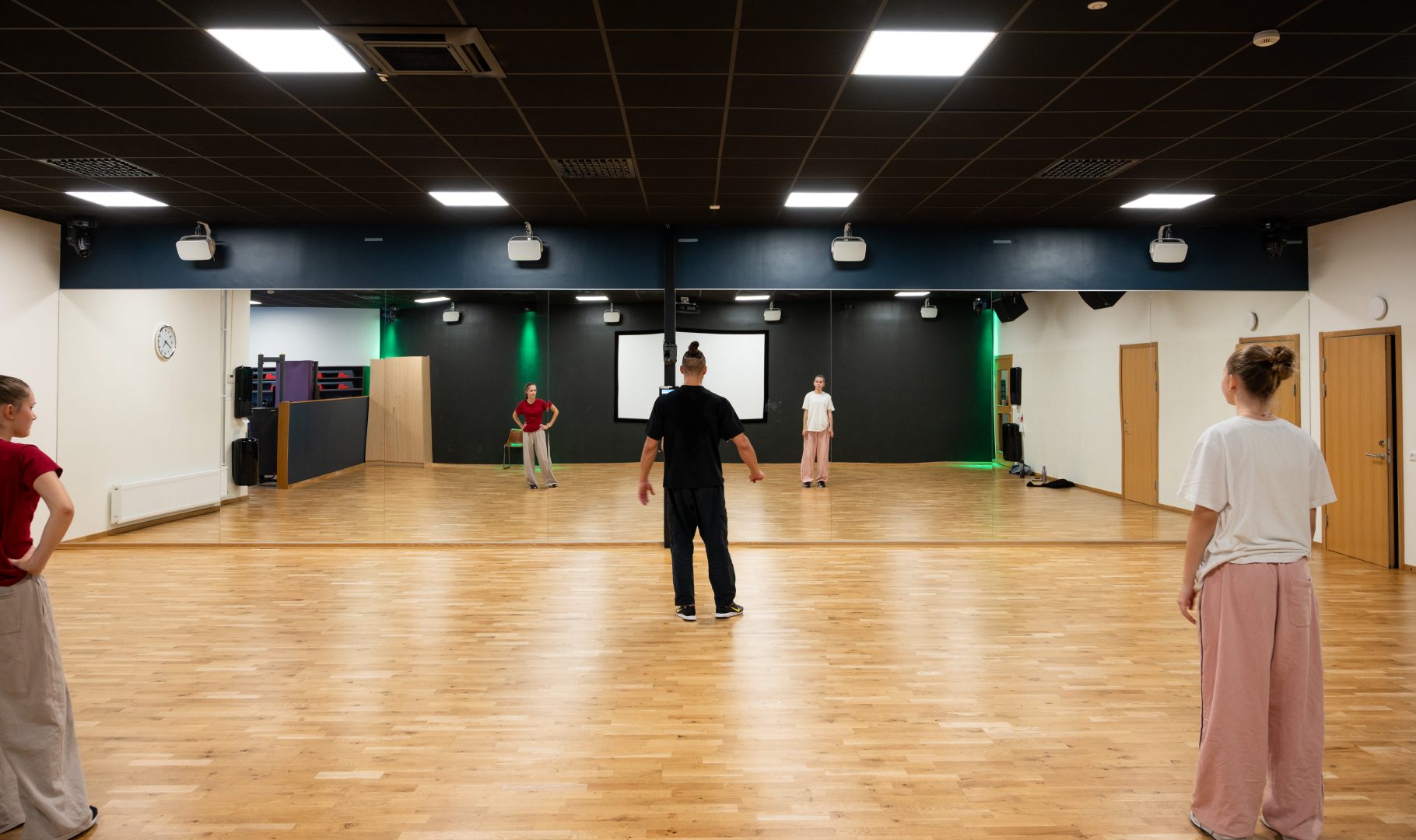 En grupp personer står i en danssal med trägolv och stora speglar längs väggen. En instruktör står i mitten med ryggen mot kameran, medan deltagarna står sprida framför honom, redo att följa instruktioner. rummet är väl upplyst med taklampor och har dörrar längs göra vägen.