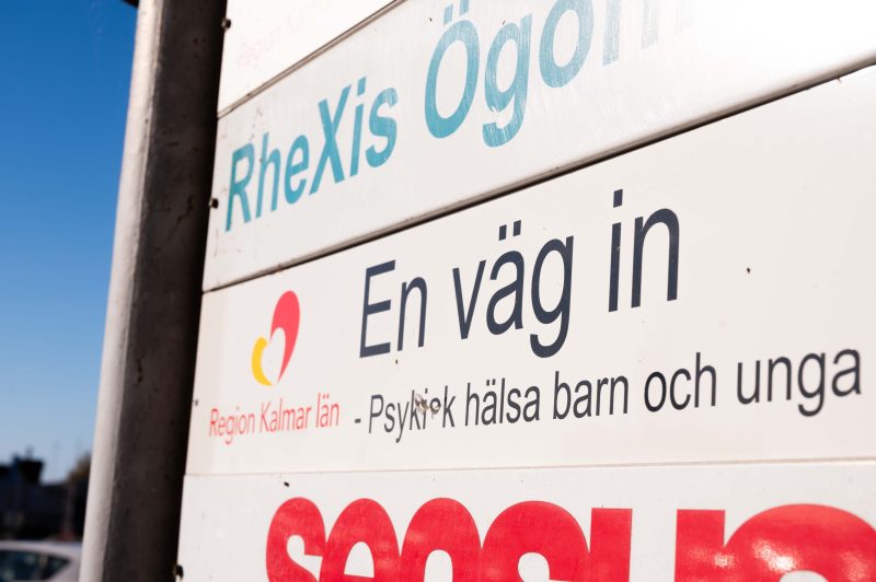 På bilden syns en vit skylt med svart text. På skylten står det "En väg in. Psykisk hälsa barn och unga." Bredvid syns en röd och gul logga, under loggan står det "Region Kalmar län". I bakgrunden syns en blå himmel.