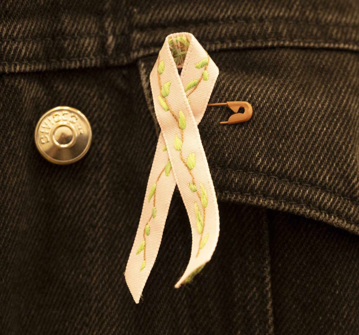 En närbild av årets rosa band. Det är en ljusrosa bandrosett med grönt broderimönster, fäst med en säkerhetsnål på en mörk jeansjacka