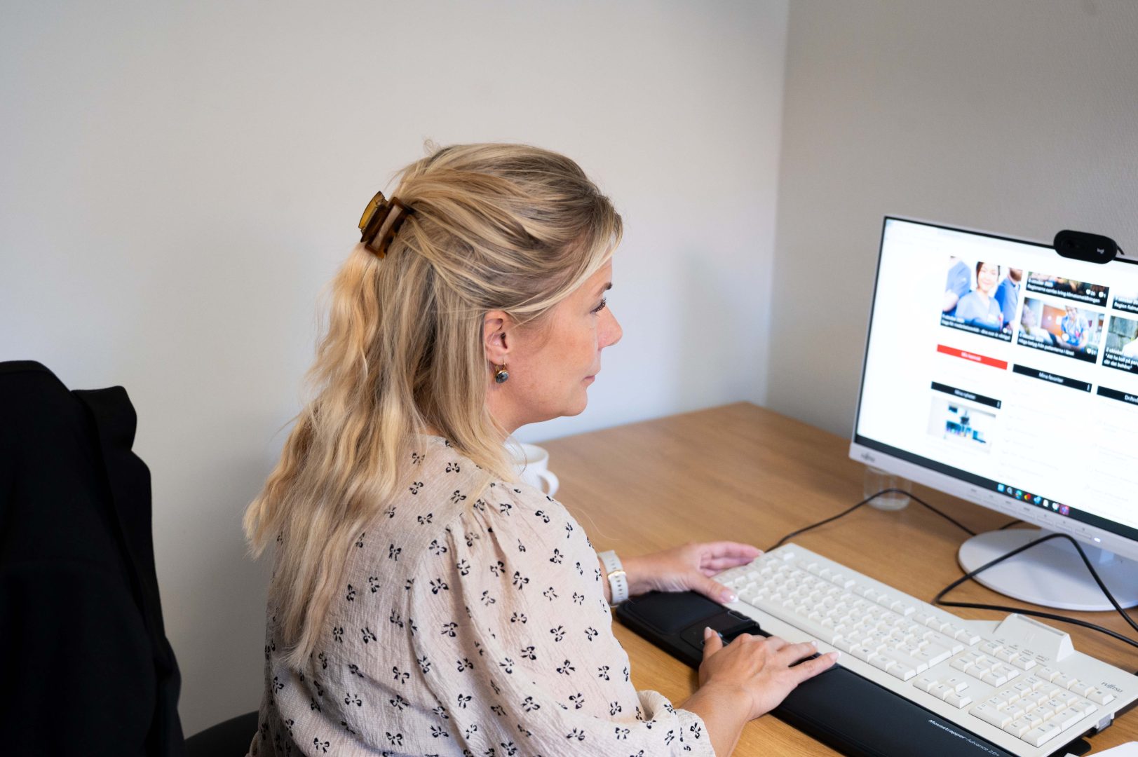 På bilden syns en kvinna som sitter bortvänd från kameran. Hon tittar på en datorskärm och har fingrarna på tangentbordet. kvinnan har en vit blus.