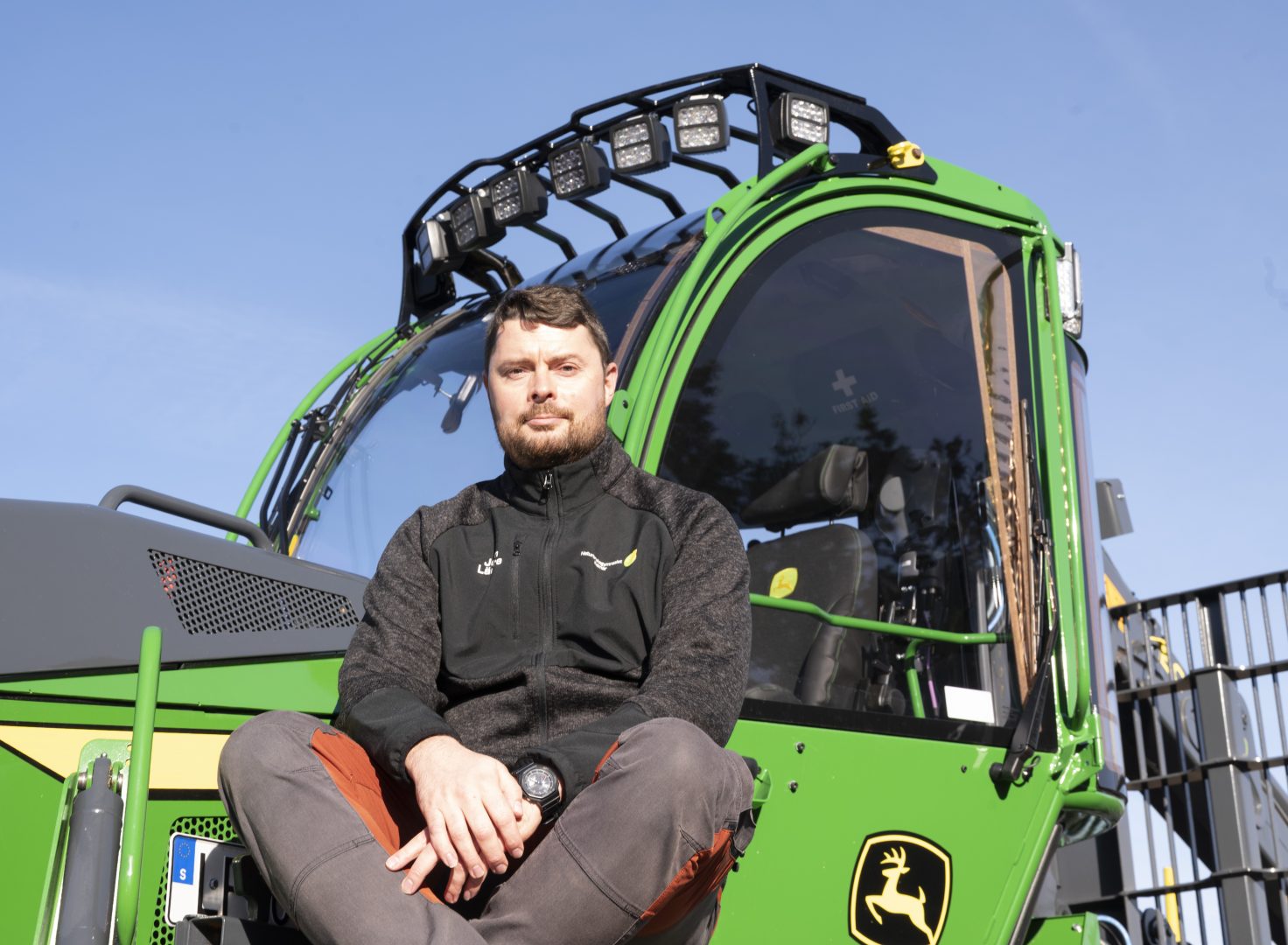 en man sitter på en grön skogsmaskin. han har på sig en svart jacka och ett par mörka arbetsbyxor.
