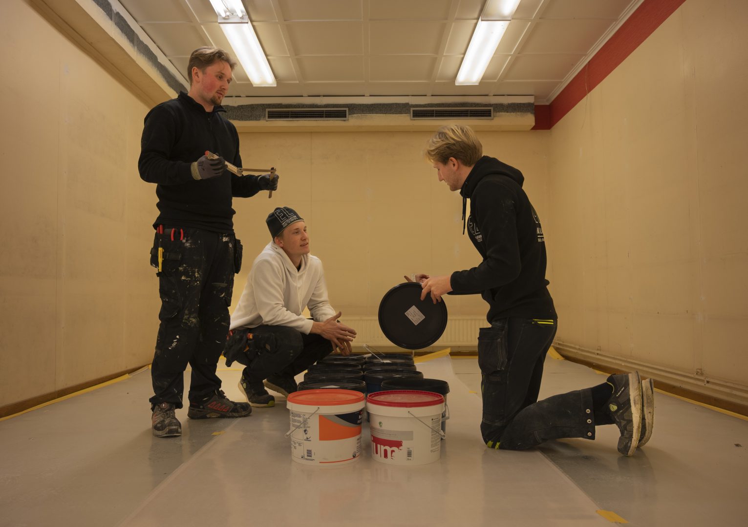 Tre killar i arbetskläder konverserar med varandra. Emellan dem står det flera burkar färg och en av killarna har öppnat en burk.