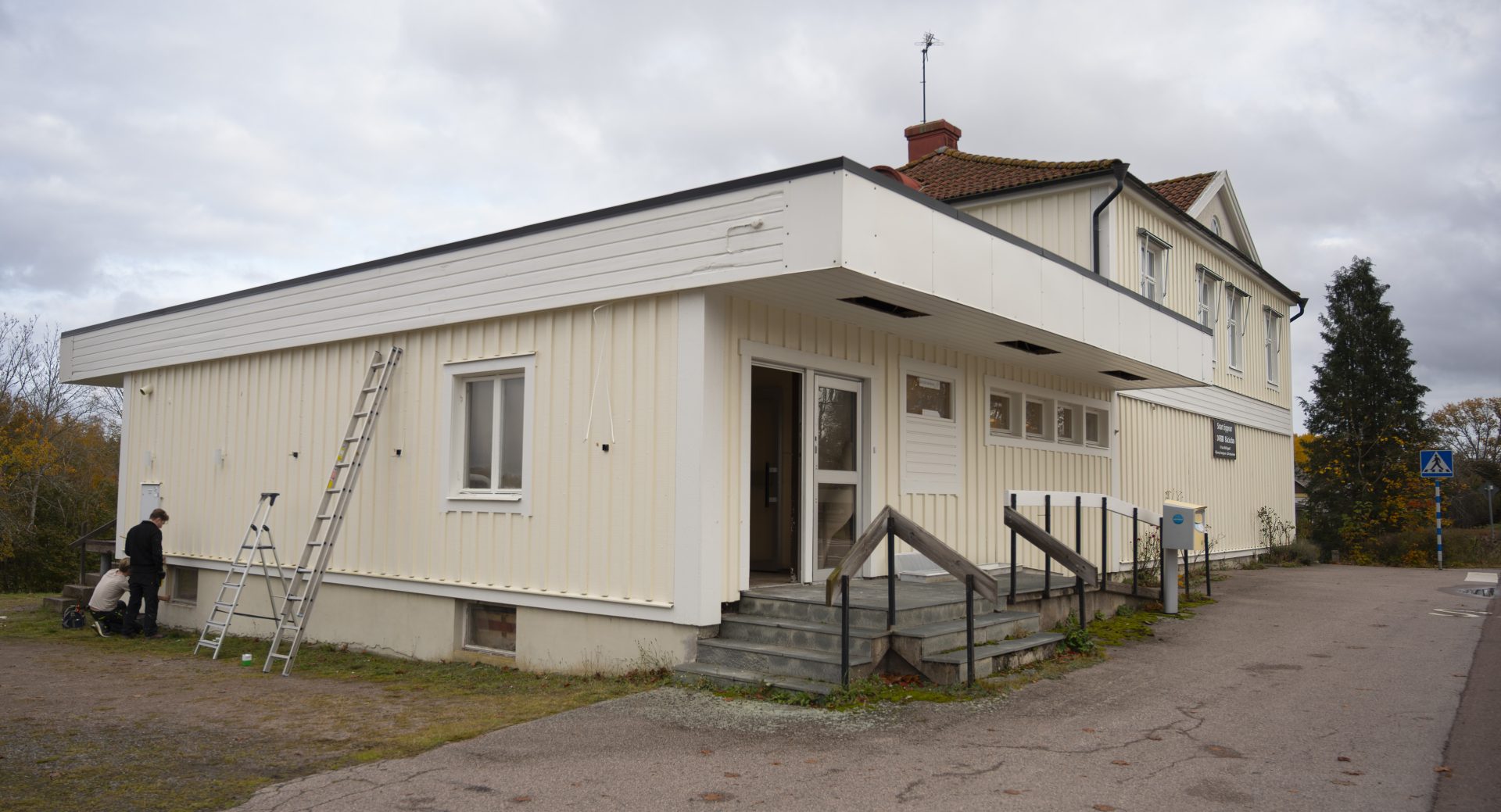 Ett stort ljusgult hus med en liten trappa upp till ingången. Himlen är grå. Det står stegar lutade mot huset och två killar står vid fasaden och arbetar.