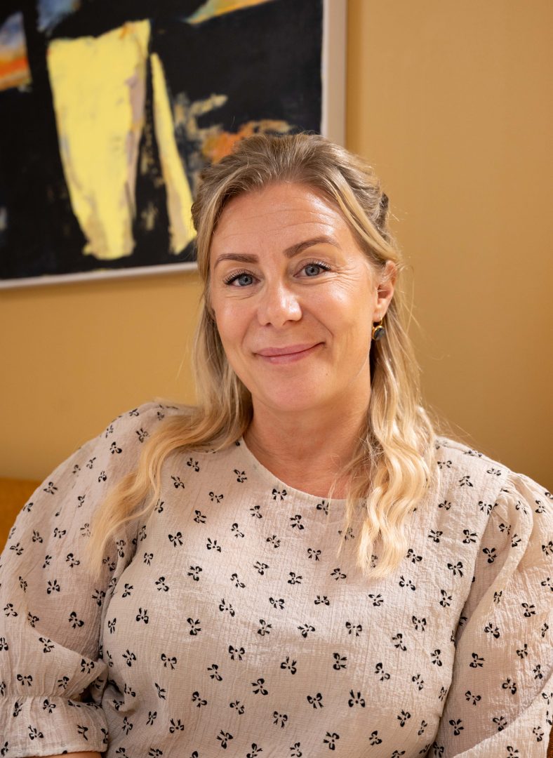 På bilden syns en ljushårig kvinna som ler mot kameran. Hon har på sig en vit blus. Bakgrunden är gul.