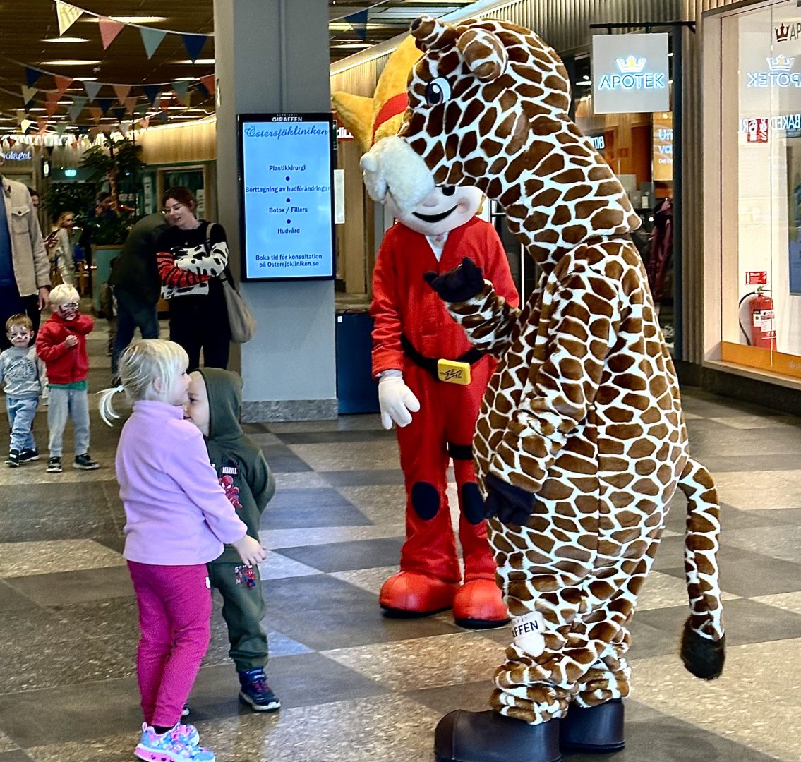Två små barn pratar med två maskotar, den ena maskoten är klädd som en giraff och den andra bär en röd dräkt med gult hår. 