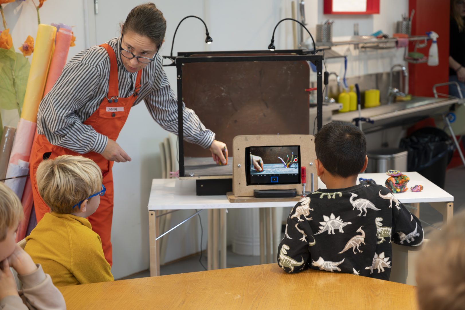 En kvinna som står framför en grupp barn, som tittar på henne och något som hon visar, i ett kreativt eller utbildande sammanhang.