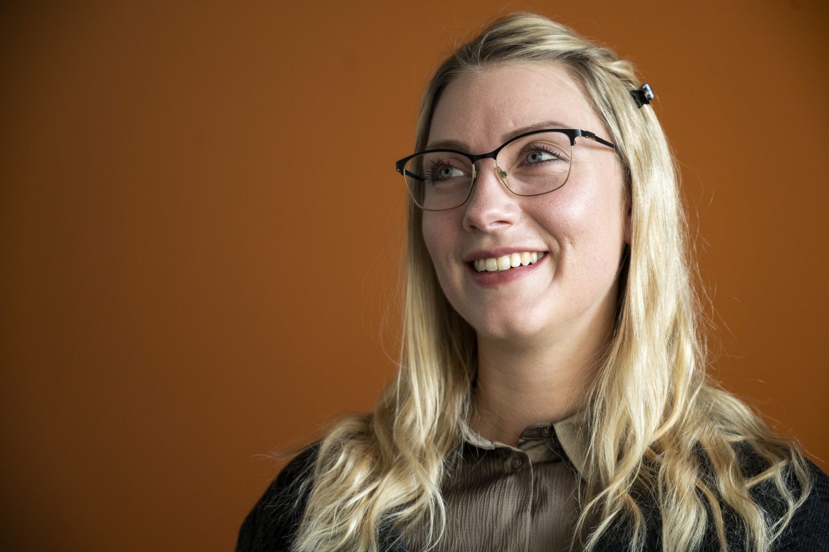 En person med långt blont hår och glasögon ler och ser uppåt. Hen bär en brun skjorta och en mörk kofta, och står framför en varm orange bakgrund som skapar en harmonisk kontrast till hårets ljusa nyans