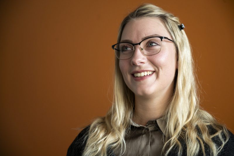 En person med långt blont hår och glasögon ler och ser uppåt. Hen bär en brun skjorta och en mörk kofta, och står framför en varm orange bakgrund som skapar en harmonisk kontrast till hårets ljusa nyans