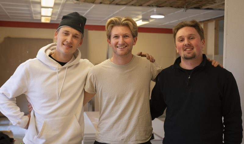 En närbild på tre killar som kollar in i kameran och ler. De håller om varandra och bär arbetskläder. En av dem har mössa på sig. De står i en stor lokal.