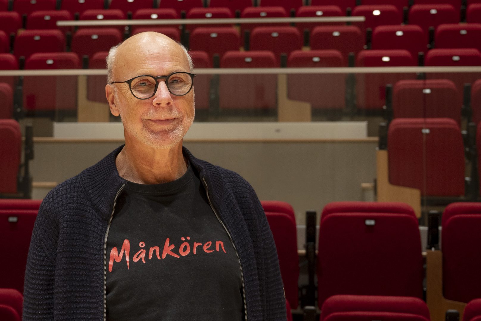 Thomas Hertzman ler mot kameran. Han bär glasögon, mörkblå kofta och en T-shirt med trycket "Månkören". Han står framför röda stolar i konserthallen i kulturhuset.