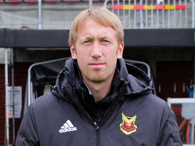Bilden visar Graham Potter som står utomhus på en idrottsanläggning. Han bär en svart jacka med ett Adidas-märke på vänster sida och en gul-röd logotyp med text på höger sida. Potter har ljust hår och tittar rakt in i kameran med neutral min. I bakgrunden syns läktare och byggnadsdelar i grått och rött.