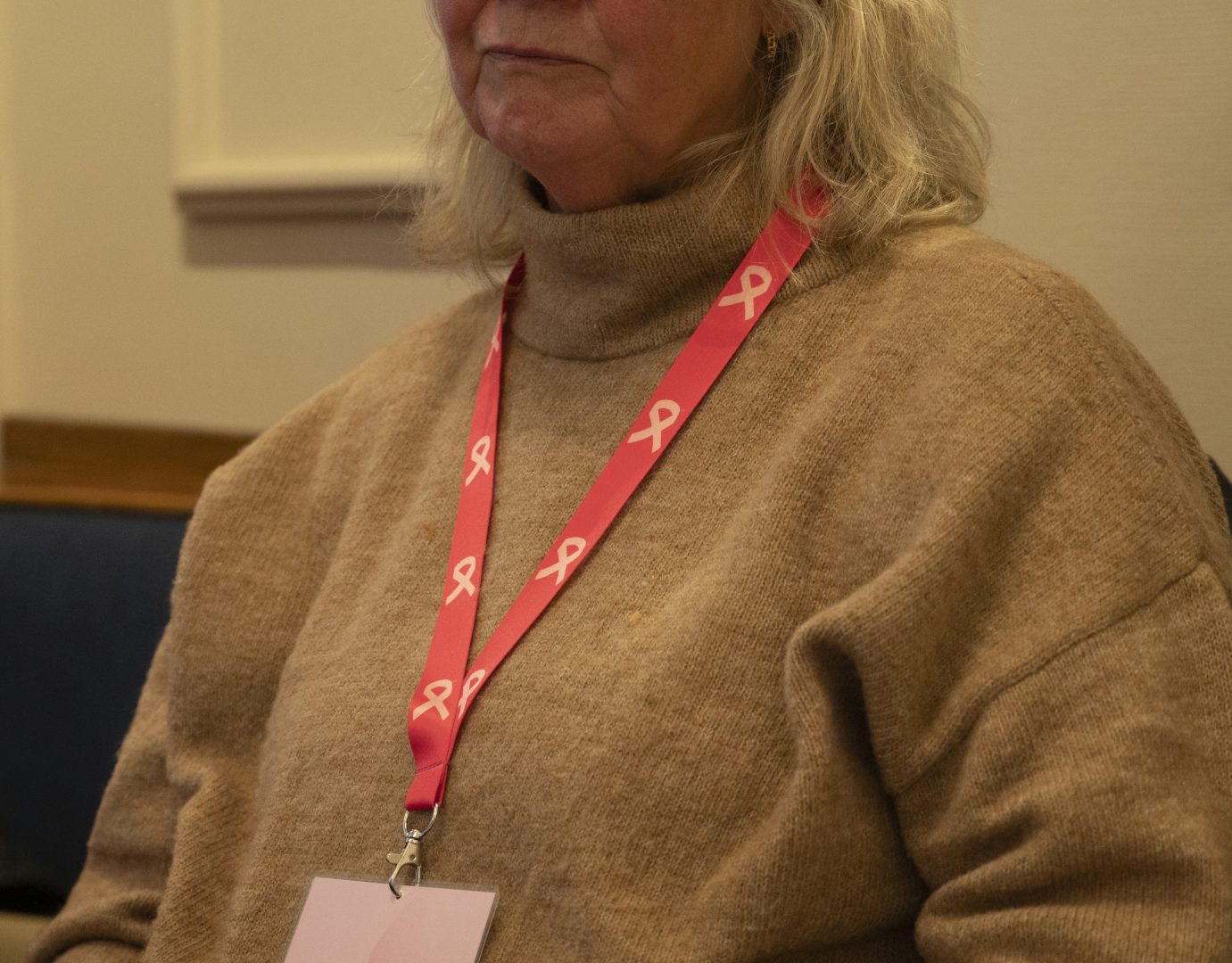 En kvinna med ljust hår bär en beige stickad tröja och ett rosa band runt halsen med vita band-symboler för bröstcancer. Bandet håller ett namnkort som hänger ned mot bröstet.