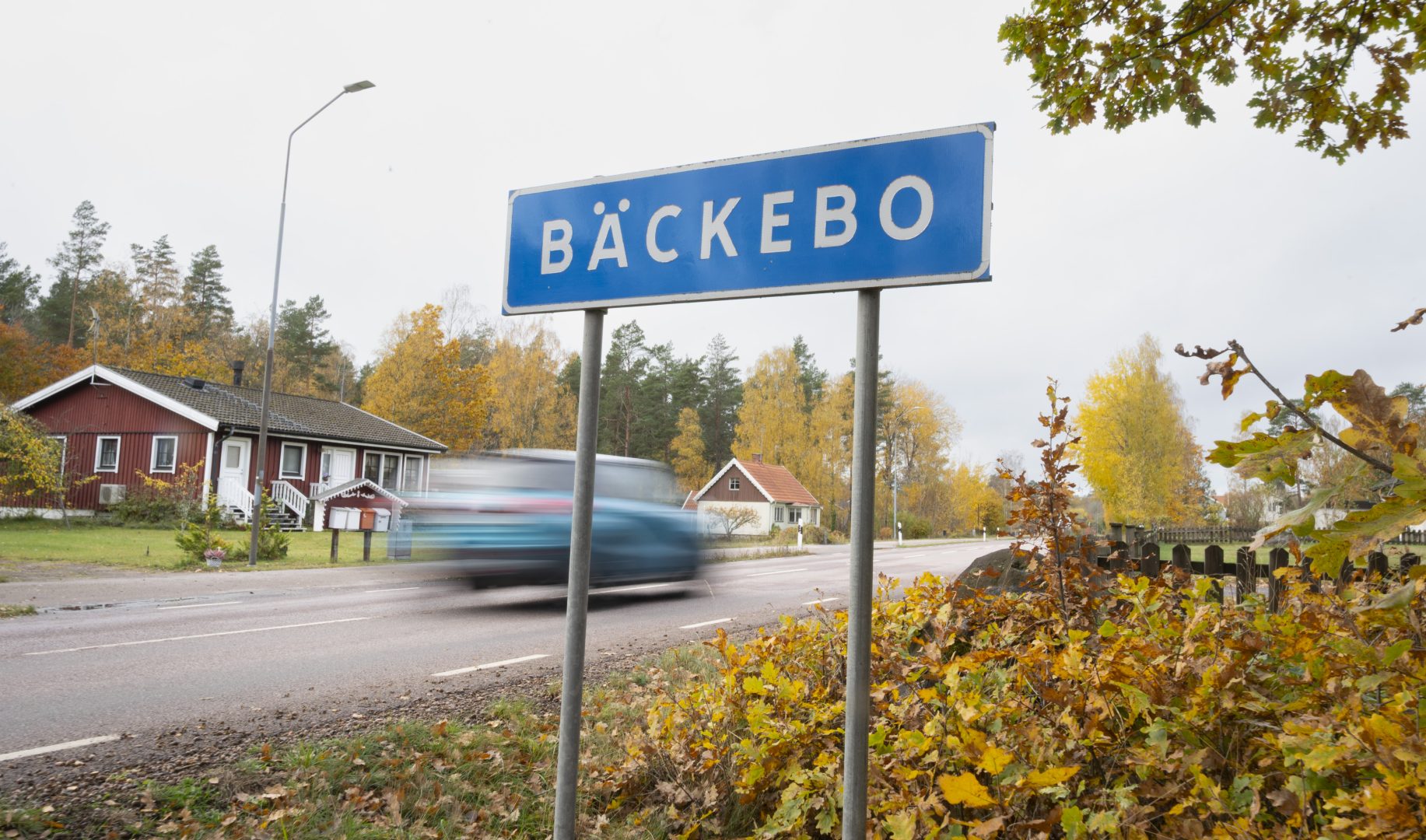 En vägskylt där det står ortsnamnet "Bäckebo". Löven runt om skylten är gula och bruna och himlen är grå. I bakgrunden syns röda hus. En blå bil kör snabbt förbi skylten, och den syns suddigt.