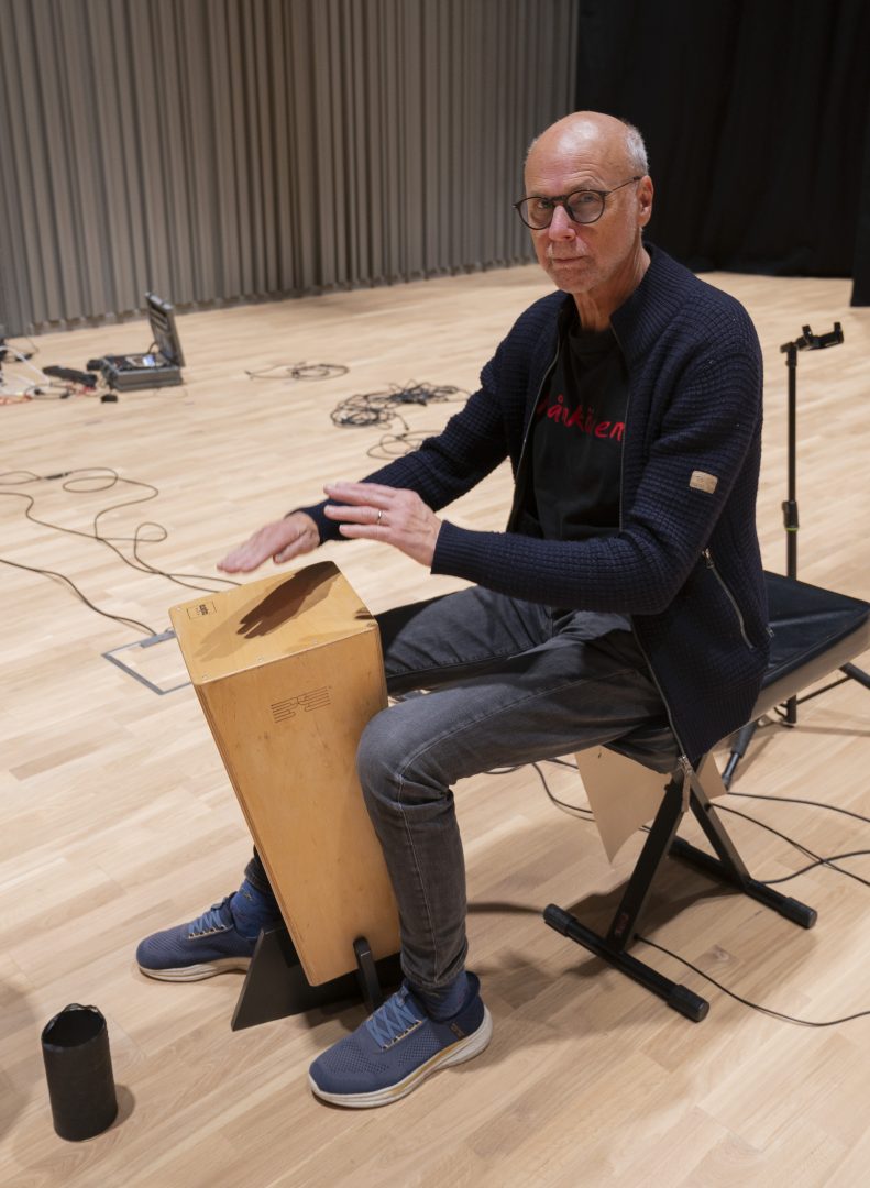 Thomas Hertzman tittar in i kameran och trummar på en canjon. Han sitter på en svart pall och det ligger sladdar på hela golvet.
