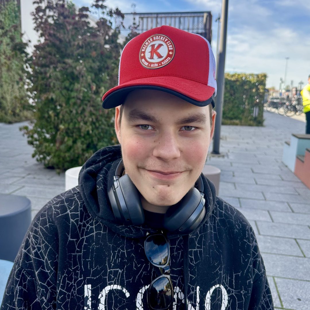 En ung kille i en röd keps med hörlurar runt halsen. Han bär en hoodie och i horden sitter ett par solglasögon. Han ler och tittar in i kameran. 