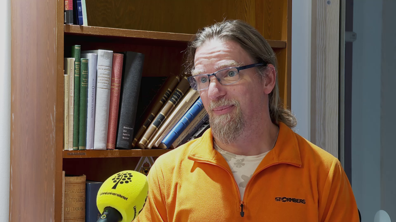 En man i orange tröja står framför en bokhylla och intervjuas.