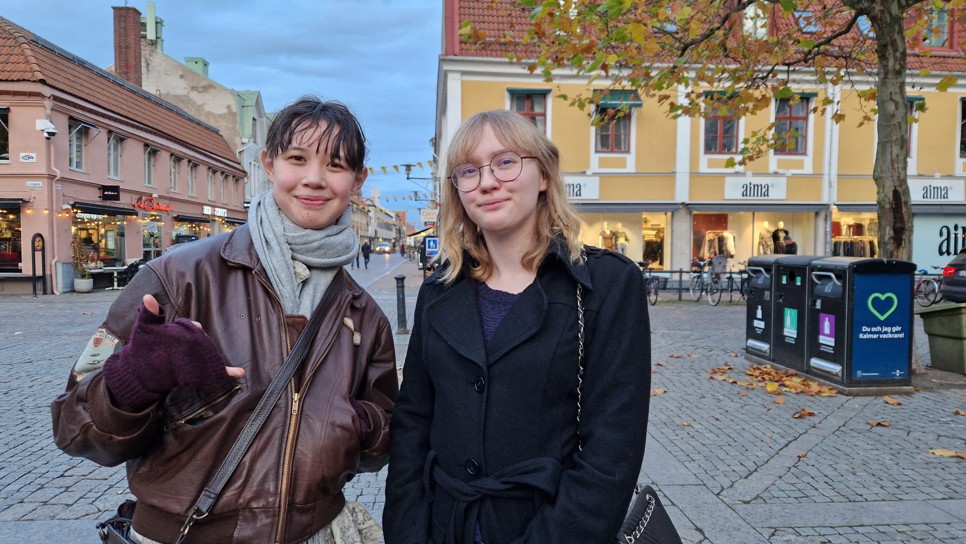 Två yngre tjejer står och kollar in i kamran. Den ena har brun läder jacka med grå sjal på sig. Den andra har en svart kappa och glasögon på sig. I bakgrunden står ett flera byggnader med affärer.