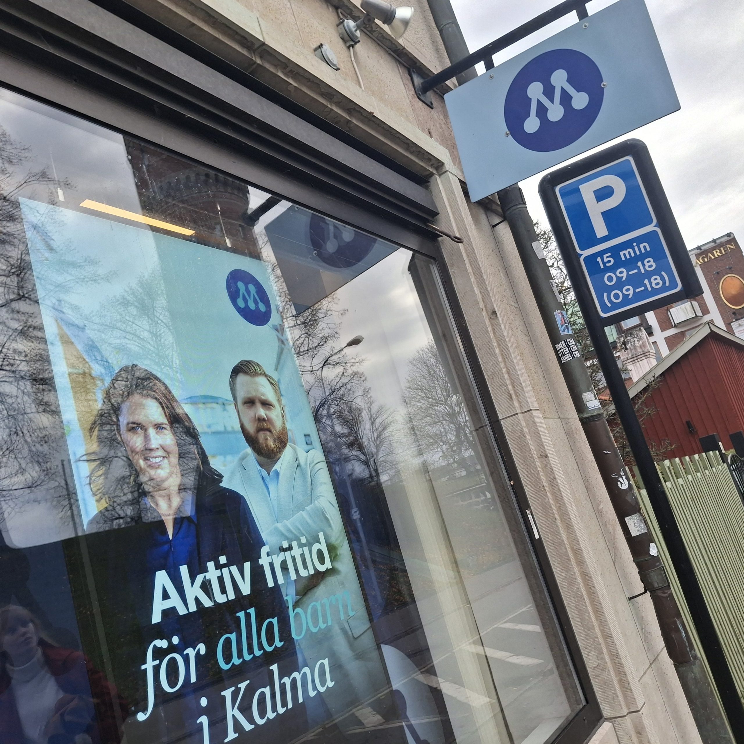 En byggnadsfasad med ett stort fönster där en politisk kampanjaffisch från Moderaterna är uppsatt. Affischen visar två personer med suddade ansikten och texten "Aktiv fritid för alla barn i Kalma". Ovanför fönstret sitter en skylt med Moderaternas logotyp – ett vitt "M" mot blå bakgrund. Till höger om byggnaden står en parkeringsskylt som anger 15 minuters parkering mellan kl. 09–18. I bakgrunden syns trottoar, andra byggnader och några gående personer.